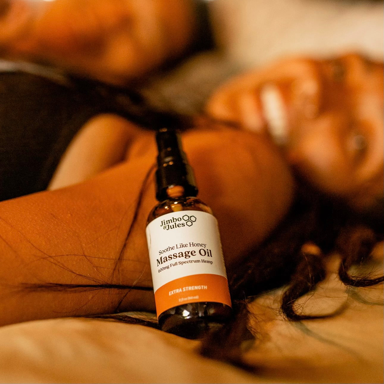 A smiling woman laying in her bed with Jimbo & Jules extra strength full spectrum CBD massage oil resting on her shoulder.