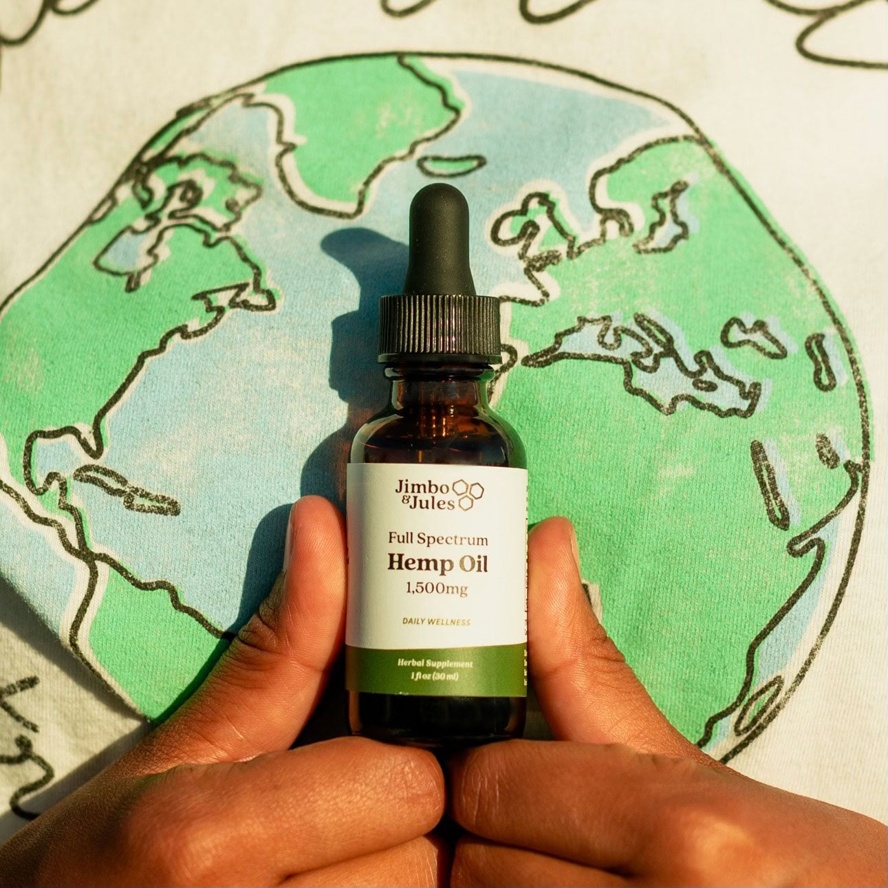 Two hands holding Jimbo & Jules 1,500mg Full Spectrum Hemp Oil in front of a shirt with a drawing of the Earth on it.