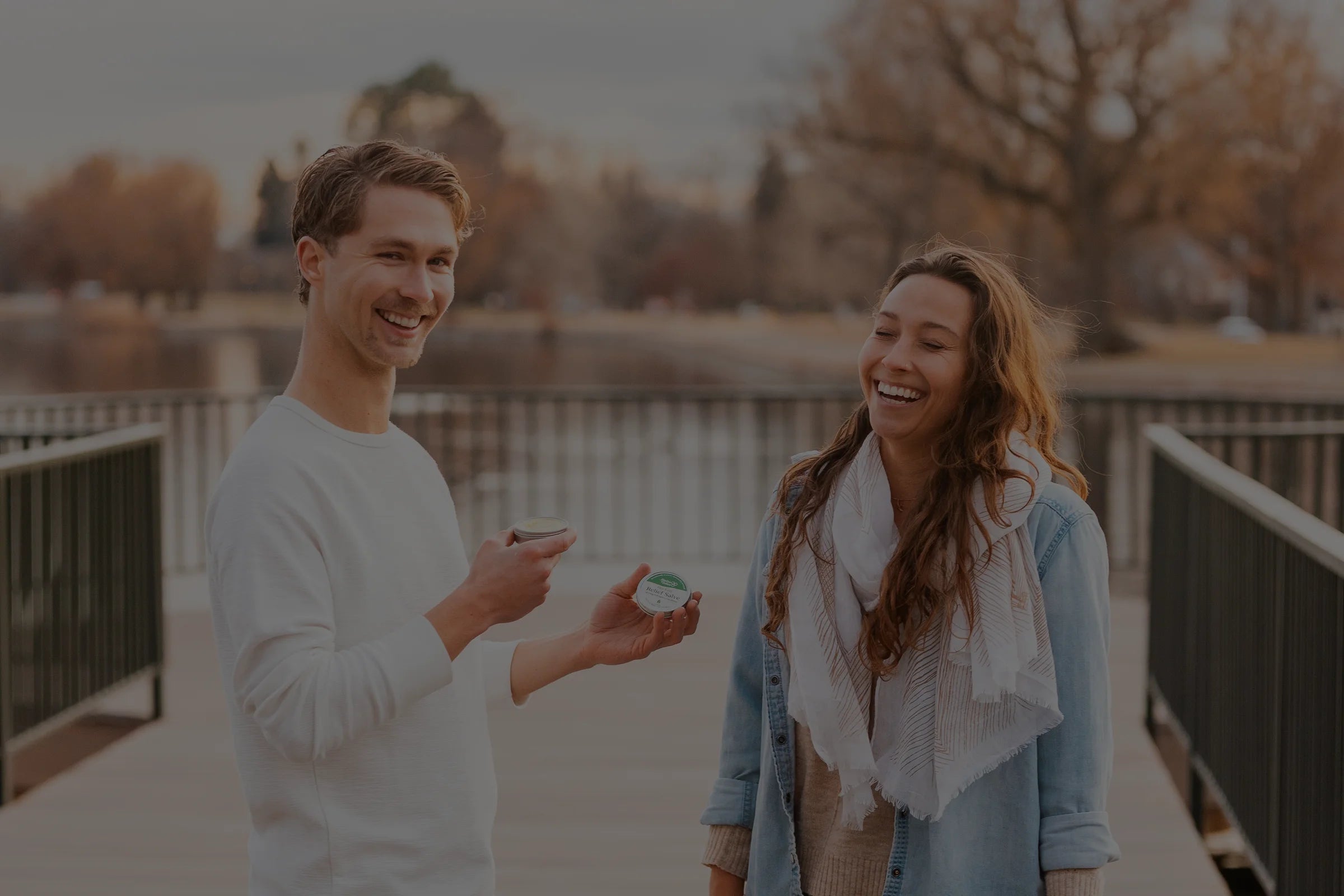 Smiling man showing a small tin of balm or cream to a woman while they stand on a wooden bridge near a lake, with autumn trees in the background.