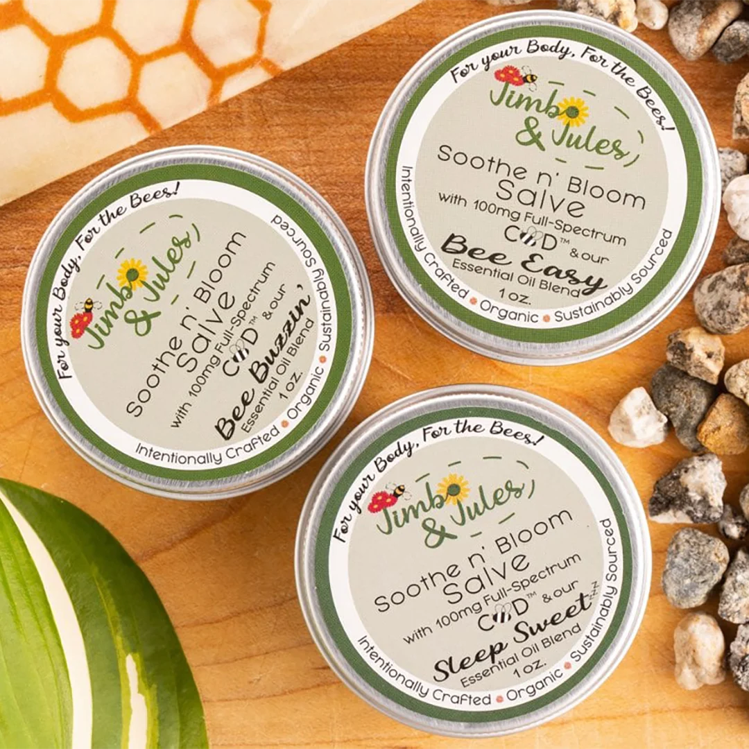 Three small tins of Jimbo & Jules CBD salve with bee-themed labels on a wooden surface.